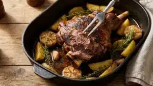 Épaule d'agneau rôtie au four dans un plat en fonte avec pommes de terre, ail et thym, une fourchette détachant la viande fondante de l'os